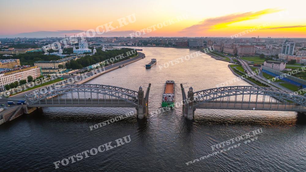 Фотография демонстрирует Большеохтинский мост через реку Неву в Санкт-Петербурге. Мост разведён, и сквозь его пролёты проходит буксируемое судно, создавая динамичную и живописную городскую сцену. На заднем плане виднеются городские кварталы и плавно освещаемое закатным солнцем небо. Этот кадр может использоваться в различных публикациях, посвящённых городскому транспорту и инфраструктуре, в туристических материалах, а также как часть визуального контента для иллюстрации статей о Санкт-Петербурге.